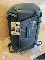 Front side view of the black THULE RoadTrip Boot Backpack with tag attached, showing the large zippered main compartment and logo.