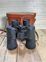 Front view of black Bushnell binoculars with the brown leather case in the background on a wooden surface.