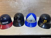 Four plastic collectible baseball helmets from left to right: Chicago White Sox, Colorado Rockies, Toronto Blue Jays, and Pittsburgh Pirates, showing team logos and condition.