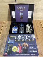 Opened box showing digital massage therapy device components: controller, large electrode pad, white wires, user manual, packaging box with product information and graphics.