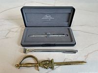 Fisher Space Pen in fitted gray foam case, below are a brass sword-shaped letter opener and an extendable metal back scratcher with a claw-shaped end.