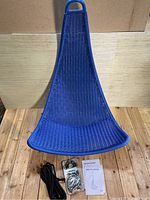 Blue woven hanging chair with a rattan-style design, black braided rope, sealed mounting hardware, and instruction manual on wooden floor.