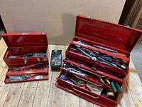 Two red metal toolboxes on wooden surface, one large with multiple trays open showing hand tools and one smaller with tools inside