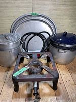 Photo shows three large metal trays standing vertically in the background, two large pots on either side, and a metal gas burner in front with attached hose.