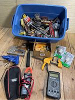Blue plastic bin filled with assorted small tools and hardware including clamps, tape measures, light bulbs, a multimeter, hand tools, a staple gun, and electrical testing equipment.