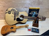 Natural wood acoustic guitar with black dragon graphic, smaller wooden guitar, metronome, Tragically Hip sheet music, Yamaha tuner, and red picks arranged on wooden surface.