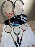 Five rackets arranged with two modern tennis rackets standing against case and three wooden rackets laid flat, with can of Wilson tennis balls and Babolat case behind them.