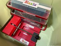 Red Sizzix manual die-cutting machine with attached handle and two plastic cases stacked containing several blue and gray die-cutting dies.