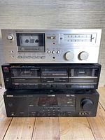 Front view of three stereo components stacked vertically on a wooden surface. Top is Ken Tech silver dual cassette deck model 4000, middle is black Scott stereo dual cassette deck DS2000, bottom is black Yamaha RX-400 receiver.