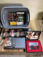 Top down view of grey bin filled with assorted VHS tapes, DVDs stacked behind, DVD/CD player with remote in front.