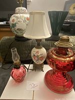 Photo showing all five lamps together on a white surface including cranberry glass oil lamp, mini cranberry glass electric lamp, hand-painted floral lamp with shade, green glass Art Deco style lamp, and Chinese Famille Rose ginger pot lamp.