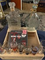 Lot arranged in wooden box with two clear cut glass bottles (one Old Forester), Rabbit Wine Tool Kit in branded packaging, and assorted souvenir shot glasses including a boot shaped one