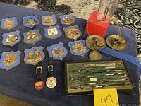 Full view showing 12 cast iron plaques arranged on cloth, engineering drafting tool set in wooden box labeled, apothecary scale with brass weights, and motorcycle keychains/tags.