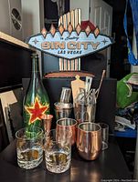 Photo showing the full set of barware including glassware, cocktail shakers, bar utensils, large Heineken bottle, and Sin City sign.