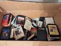 Open cardboard box containing over 75 assorted 8 track tapes. Various colors and labels visible, items loosely stacked inside box.