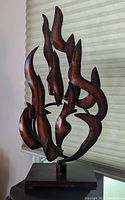 Photo showing a dark reddish, orange, and brown toned Mid-Century Modern metal flame sculpture on a rectangular base against a window blind background.