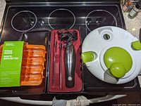 Photo of electric handheld carver in red case, orange kebab maker mold in plastic packaging, and white-green salad spinner on stove