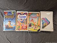 Four Disney VHS tapes laid side by side on a fabric surface, showing front covers including The Little Mermaid, Snow White, Bambi, and a fourth tape.