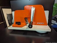 Front and side view of orange hand crank meat and bread slicer on white base with suction cup.