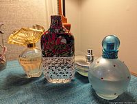 Four perfume bottles photographed on a blue fabric surface including BCBG Max Azria Bon Chic, FCUK Pulse with graffiti design, Britney Spears Circus with frosted round bottle and blue cap, and Nautica My Voyage with silver flat bottle.