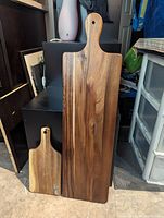 Photo showing two wooden charcuterie boards, one large and one small, standing upright with handles and hanging holes, natural wood grain visible.