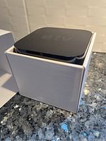 Apple TV device placed on its white box, showing top and side view with embossed 'tv' logo.