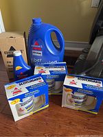 Blue bottle of Bissell fiber cleaning solution, smaller bottle of Bissell cleaning fluid, and three boxes of Scotchgard protection on wood floor near cardboard box.