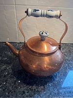 Side view of rounded brass-colored tea pot with arched ceramic handle decorated with blue floral pattern, ceramic knob lid, placed on a granite countertop with textured white tile backdrop.