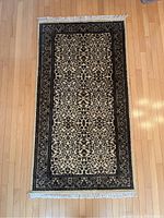 Full view of the 3x5.5 ft rectangular carpet with central floral pattern, dark decorative border, and white fringe tassels at both ends, placed on a wooden floor.