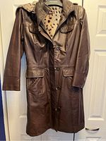 Front view of the brown leather jacket with button closure, flap pockets, and attached faux fur hood hanging on a door.