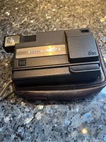Front view of Kodak Tele Disc camera resting on brown pouch, showing the camera body and flash on left.