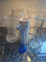 Overall view of seven flower vases in various sizes and colors including clear glass, white ceramic, and blue glass.