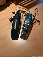 Side-by-side view of two hand vacuums on floor: black Eureka plug-in and blue Hoover rechargeable with cords.
