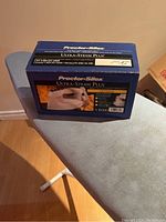 Photo of the boxed Proctor Silex Ultra-Steam Plus steam iron sitting on top of a gray ironing board with white collapsible legs on a wooden floor.