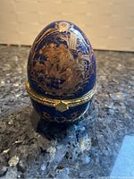 Underside view showing closed dark blue egg with intricate gold floral and scroll pattern, gold-tone hinge and rim.