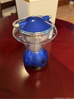 Blue mini lantern, dome-shaped top, clear plastic surrounding and metal handle, sitting on a table.