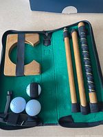 Top-down view showing wooden putter cup, disassembled wooden putter, practice balls, and small putter head in green felt-lined case