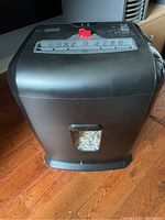 Front view of the black Staples paper shredder showing the body, viewing window for shredded paper level, and hardwood floor.