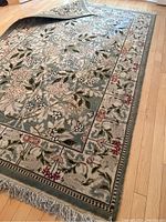 Photo showing the full rectangular rug laid out on a wooden floor with visible floral pattern and fringe on the edges, partially folded to show rug's thickness and material.