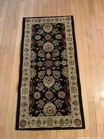 Full view of one plush decorative rug with floral motifs, black central field and cream border, laid on hardwood floor.