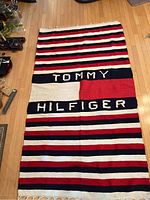 Full view of the Tommy Hilfiger striped blanket laid flat on a wooden floor, showing the brand name in large letters and the color pattern.