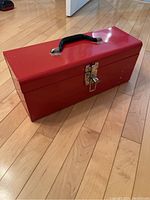 Closed red metal 16 inch toolbox with black handle and metal latch on wooden floor.