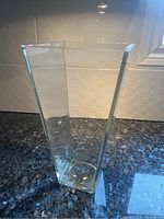 Clear glass vase photographed on a dark countertop with white tiled backsplash, showing the full vase height and square shape.