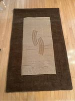 Full view of brown handmade Indian wool carpet on wooden floor showing central design and border pattern.