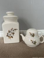 Full view of large ceramic canister next to stacked teapot and cup, all cream colored with floral decoration.