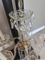 Close-up photo of the top section of the chandelier showing the clear glass circular tray and hanging crystal droplets attached to metal hooks, and the glass central column with metal fittings.