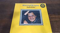 Front cover of Harry Chapin Heads & Tales vinyl LP album showing artist portrait on yellow background with black and white border.