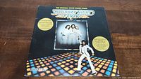 Front cover of the Saturday Night Fever soundtrack vinyl showing the iconic disco floor and characters from the movie, black background with text and yellow promo circles.