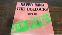 Front cover of the vinyl album titled "Never Mind The Bollocks Here's The Sex Pistols" with bright pink background and green and black text.