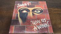 Front cover of Sweet's 'Give Us A Wink' vinyl record album showing a winking face with brick wall background and album title text.